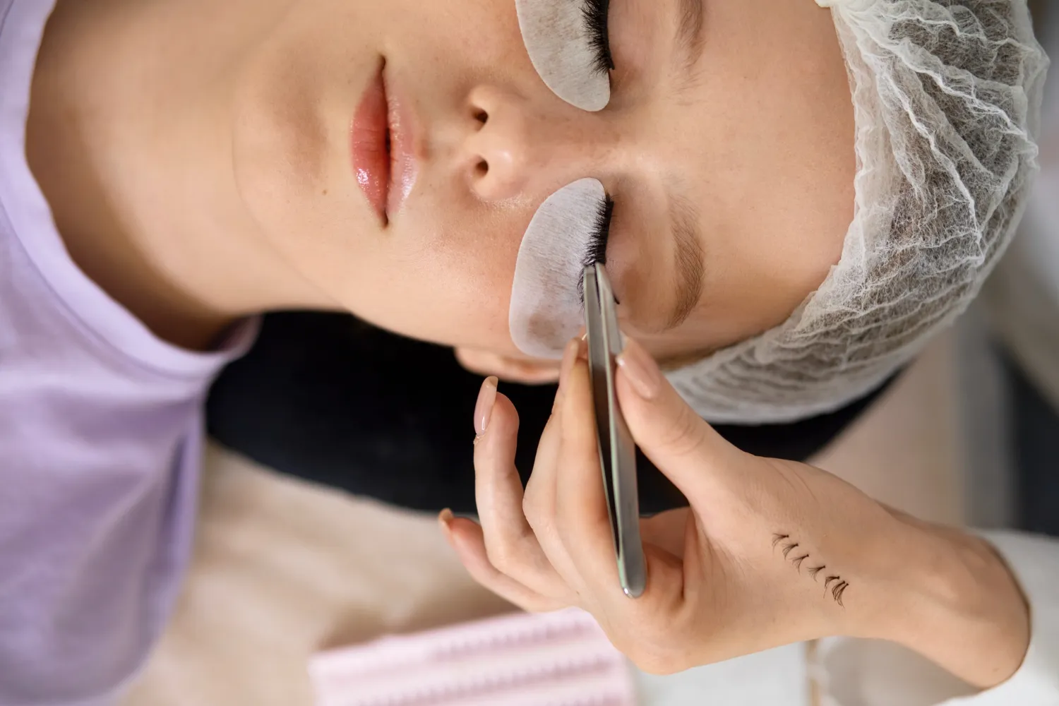 Aplicación de extensiones de pestañas en una mujer con parches protectores sobre los ojos, mientras una especialista utiliza pinzas para colocar las pestañas.