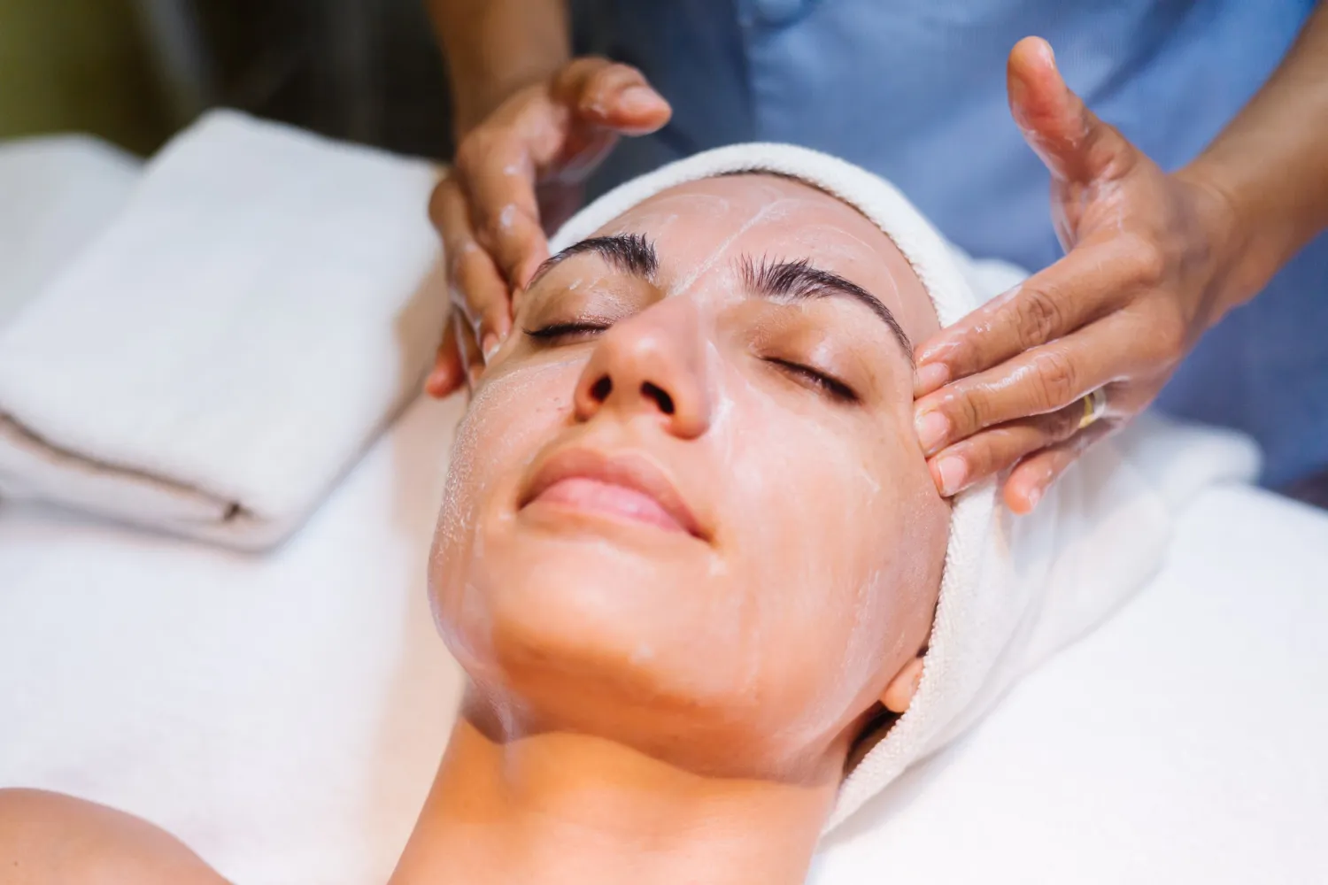 Mujer recibiendo un tratamiento facial en un spa, con toallas limpias a su alrededor, mientras un esteticista aplica un producto en su piel.