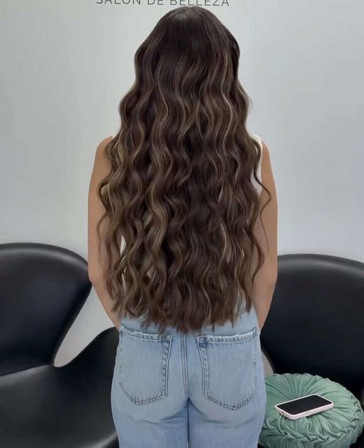 Mujer de espaldas con cabello largo y ondulado, en un salón de belleza llamado Amoracuy. El fondo muestra el logotipo del salón y muebles modernos.