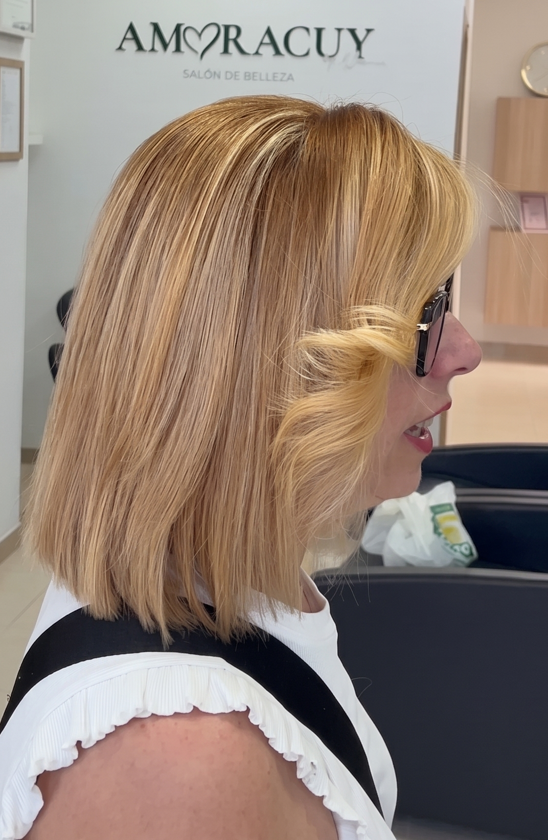 Mujer con cabello rubio y lacio, con un suave rizo en la parte frontal, en un salón de belleza llamado Amoracuy. Se le ve de perfil, con gafas oscuras y una blusa blanca con detalles en negro.