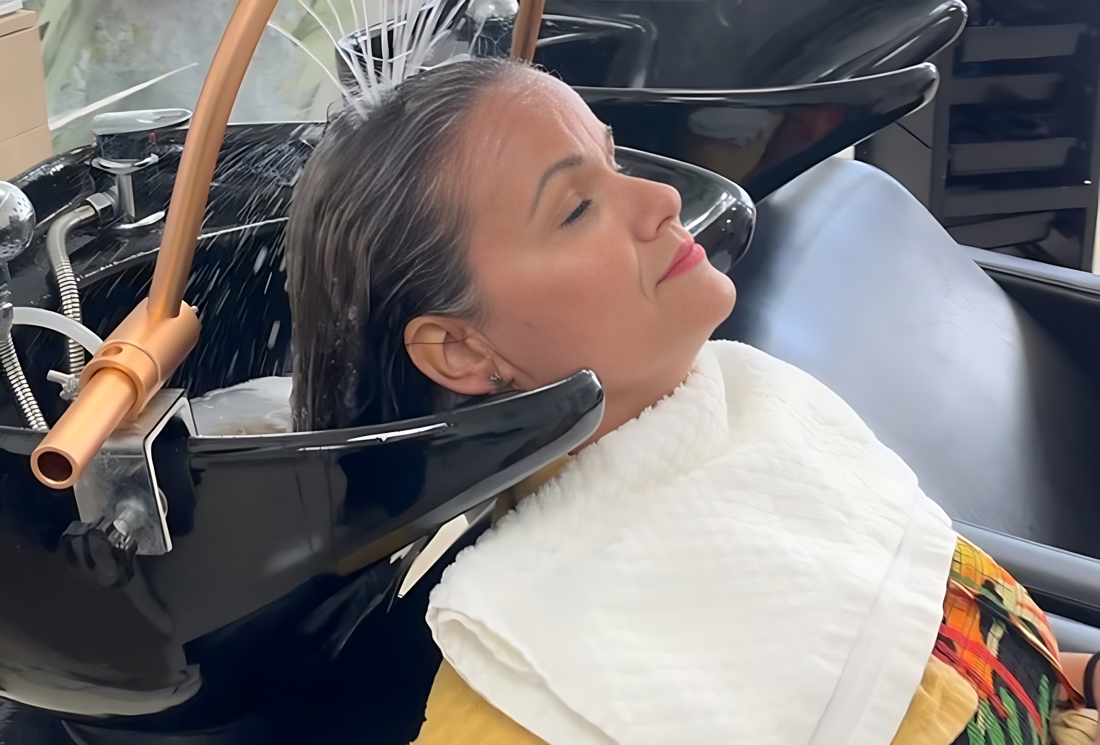 Mujer recibiendo un tratamiento capilar en un salón de belleza, con el cabello mojado y relajada, mientras el agua fluye sobre su cabeza. Viste una blusa colorida y está cubierta con una toalla blanca.